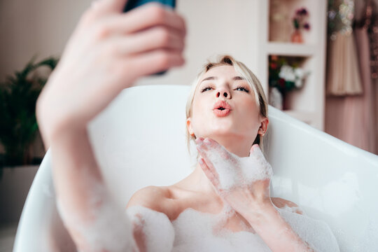 Young beautiful sexy woman having fun while lying in bathtub full of foam at home. Charming smiling model relaxing in luxury bath interior. Female holding smartphone, taking selfie photos
