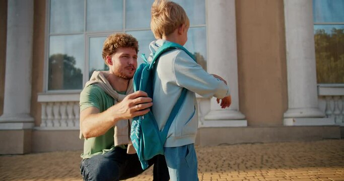 A father with curly hair in a green T-shirt sends his little blond son in a blue jacket and with a turquoise backpack to school. Little elementary school student runs to school