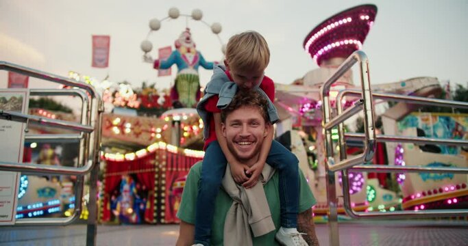 Portrait of a Happy father with curly hair in a Green T-shirt and his little blond son in a red T-shirt against the backdrop of bright lights and attractions in an amusement park