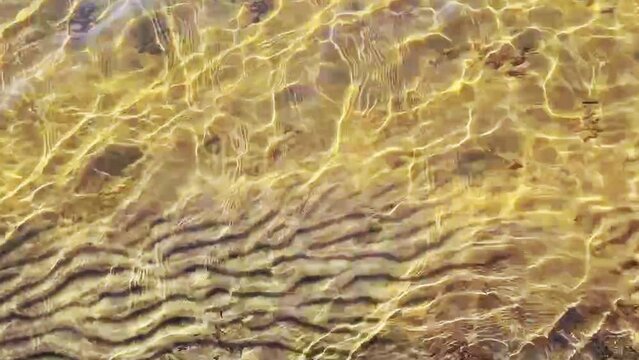 Calm lake water is flowing with small wave. Sun glitter is creating bright reflection. Textured yellow sand surface under water.