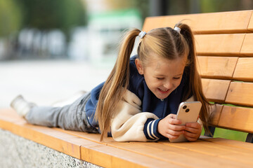 Little girl schoolgirl in park lying on a bench, holding in the hands a smartphone reads and writes to correspond in social networks, looks video on the smartphone. Rest after school in nature.
