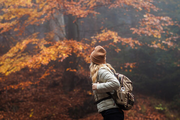Fototapeta na wymiar Woman with backpack and knit hat hiking in fog in autumn forest. Warm clothing for leisure activity in cold weather