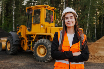 A young woman mine worker is standing in front of a large dump truck or grader. Looking at camera.