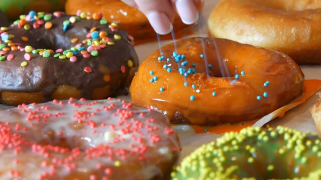 Large multi-colored fresh fried donuts in a row on a table. A beautiful donut with orange icing is sprinkled with a special colored powder for decoration of sweets