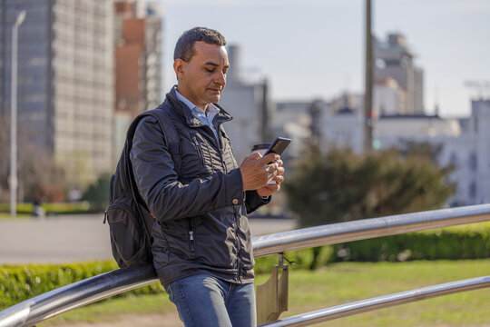 Latino man using his mobile phone and holding a disposable cup of coffee leaning on a railing in a public park with copy space.