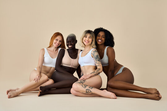 Happy diverse young women wear underwear sitting showing smooth depilated waxed skin legs on beige background. Multicultural girls, multiracial ladies beauty models group advertising body skin care.
