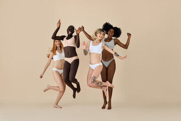 Happy funny diverse girls wearing underwear jumping on beige background. Funky pretty multicultural young women models group laughing having fun, diversity and natural body beauty, authentic shot.