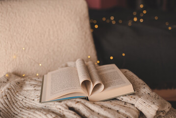 Open paper book with folded pages in heart shape on knitted clothes over Christmas lights in dark...