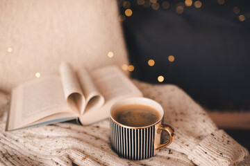 Open paper book with folded pages in heart shape and cup of coffee on knitted clothes over Christmas lights in room close up. Winter cozy home atmosphere.