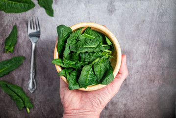 Man holding fresh spinach leaves in a wooden bowl. Background of a gray stone table, top view, cutlery, fork.