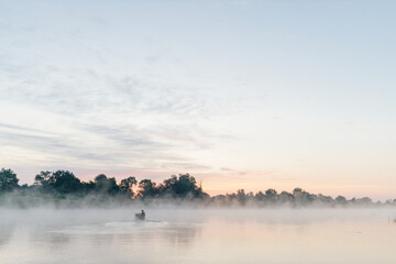 Obraz na płótnie Canvas Beautiful morning landscape with a river along which a boat floats, trees, fog and the dawn sky. Dense fog on the lake early in the morning