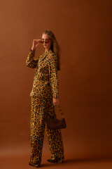 Fashionable confident woman wearing trendy leopard print satin suit blazer, trousers, sunglasses, holding small leather bag, posing in studio, on brown background. Fill-length portrait 