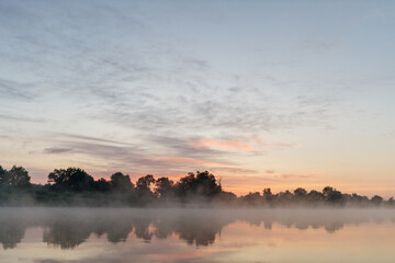 Beautiful landscape with a river and reflection, thick fog at dawn. Lake in the early morning in...