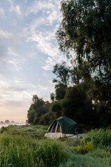 Beautiful summer landscape by the river in the early foggy morning. The tent stands among green grass and trees, on the shore of a lake. Camping. Watching dawn near a river covered in thick fog