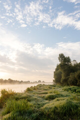 Fototapeta na wymiar Beautiful summer landscape by the river in the early foggy morning. The tent stands among green grass and trees, on the shore of a lake. Camping. Watching dawn near a river covered in thick fog