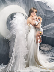 Mom and daughter in angel wings. Young woman and one-year-old baby photograph in the studio