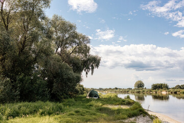 Obraz premium Beautiful summer landscape by the river on a sunny summer day. The tent stands among green grass and trees, on the shore of a lake. Camping.