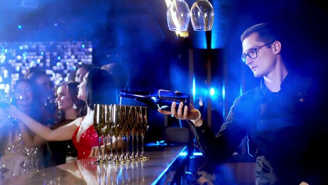 Close-up of a bar riser, the bartender pours champagne. A group of happy people at a party celebrating and drinking champagne in a club, against the backdrop of bright lights.