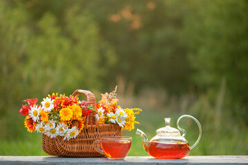 Glass Teapot and cup with herbal tea, fresh useful flowers, herbs in wicker basket on table...