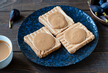 Peanut butter spread on the toasts on dark blue plate, butter in a bowl, figs over wooden background