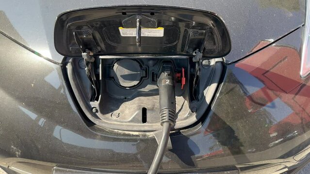 A black electric car is being charged at a charging station with a cable inserted into the charging socket in the middle of the hood of the car on a sunny day. EV charging station for an electric car