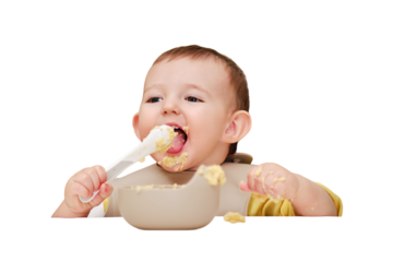 Happy toddler baby boy learns to eat porridge himself with a spoon while sitting in a child chair, isolated on a white background. Smiling child eats with his hands and a spoon, kid aged one year