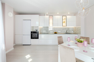 A modern living room combined with a kitchen, in light colors. Dining table with pink tableware