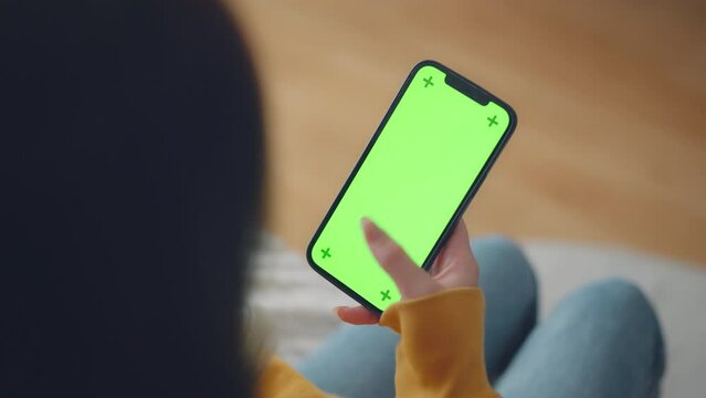 Hand of woman holding mobile smartphone and swipes photos or pictures with blank green screen mock up while sitting on a sofa at home, Chroma key mock-up on smartphone in hand