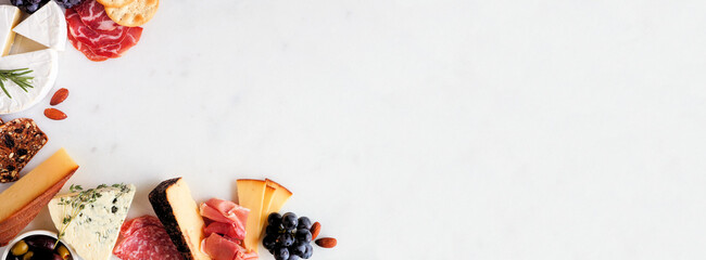 Selection of charcuterie cheeses, meats and appetizers. Above view corner border on a white marble background with copy space.