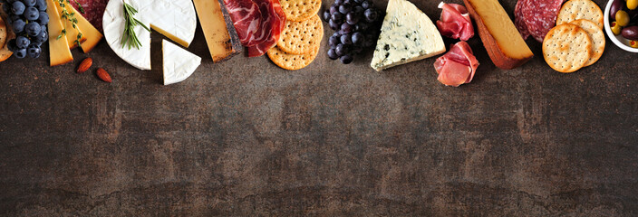 Variety of charcuterie cheeses, meats and appetizers. Overhead view top border on a dark stone background with copy space.