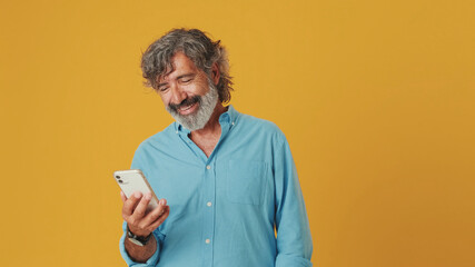 Happy elderly grey-haired bearded man wears blue shirt, uses mobile phone, having fun with mobile device, isolated on orange background in studio