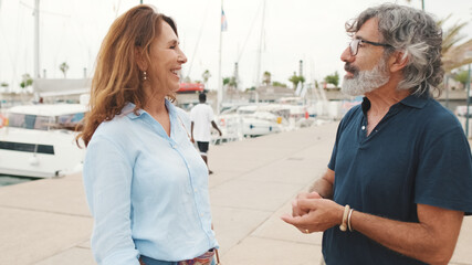 Obraz premium Smiling middle aged couple talking while standing in marina on yacht background