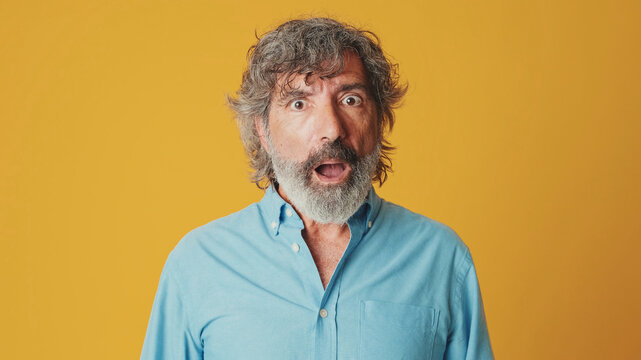 Close-up of an elderly grey-haired bearded man wearing a blue shirt looking at the camera with surprise, wow effect isolated on orange background in studio