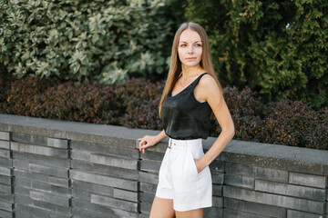 Portrait of a girl with long blond hair, wearing a black top, white shorts, poses against a gray wall. Young woman walking through a park with modern architecture and landscape design
