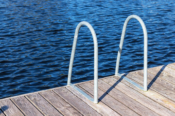 Metal dock ladder on wooden pear, blue water. Sea, ocean, river or lake. Summer swimming time.

