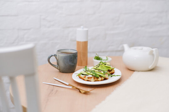 Breakfast on wooden table:zucchini waffles with cottage cheese, avakado and arugula in a white plate, cup of tea. Top view.