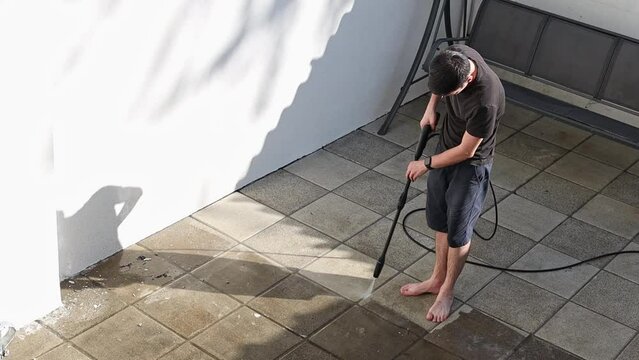 One young Caucasian brunette man in dark clothes washes dirty garden tiles with a Karcher on a summer evening in the small backyard of his house, close-up view from above.