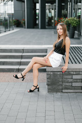 Portrait of a girl with long blond hair in a black top and white shorts posing against the backdrop of modern architecture. Young woman sitting in a park with modern architecture and landscape design