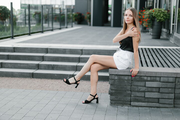 Portrait of a girl with long blond hair in a black top and white shorts posing against the backdrop of modern architecture. Young woman sitting in a park with modern architecture and landscape design