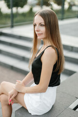 Fototapeta premium Portrait of a girl with long blond hair in a black top and white shorts posing against the backdrop of modern architecture. Young woman sitting in a park with modern architecture and landscape design