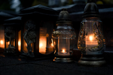 Decorative colored-glass grave candles glowing with warm candlelight on a tombstone at dusk —...
