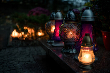 Decorative colored-glass grave candles glowing with warm candlelight on a tombstone at dusk —...