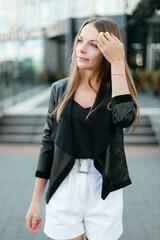 Fototapeta premium A girl with long blond hair, wearing a black top, white shorts, poses against a background of a gray wall. Young woman walking through a park with modern architecture and landscape design