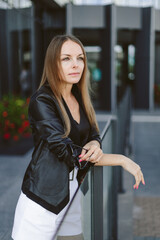 A girl with long blond hair, wearing a black top, black jacket and white shorts, poses against the backdrop of modern architecture. Young woman walking in a park with landscape design