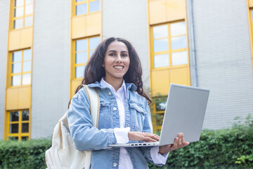 Smiling young Eastern girl stands in front of a school building or university campus holding laptop in her hands, dressed in a denim jacket and a backpack on her shoulders. Online education treatment.