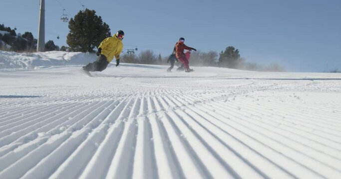 Group of people snowboarding on ski resort slope, snow spraying into the camera. Snowboarders carving on the fresh groomed corduroy slope during vacation on a sunny winter day. Slow motion