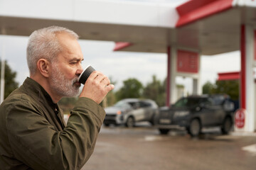 A grown man drinks coffee from a paper cup at a gas station while his car fills up with gasoline.