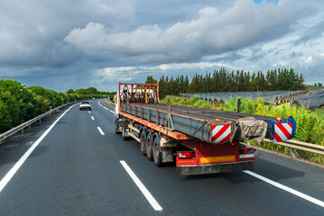 Truck loaded with excessively long iron beams, with specific signage, plates with red and white...