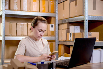 Fototapeta na wymiar Young entrepreneur working at home small business checking internet orders on smartphone and preparing boxed items for delivery to customers.
