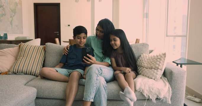 Loving Indian mother spend time with cute little son and daughter sit on couch using modern smartphone, make videocall, enjoy new cool mobile app having fun on internet. Technologies, family pastime